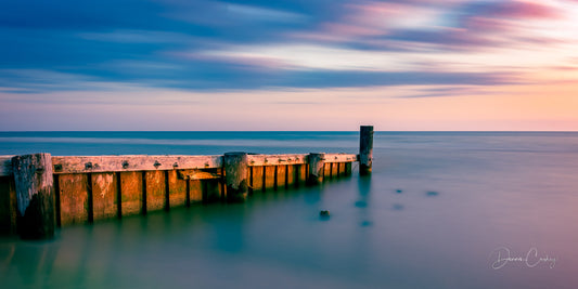 A serene sunset over a weathered jetty with soft pastel hues reflected on the water, capturing the tranquility of a summer evening.